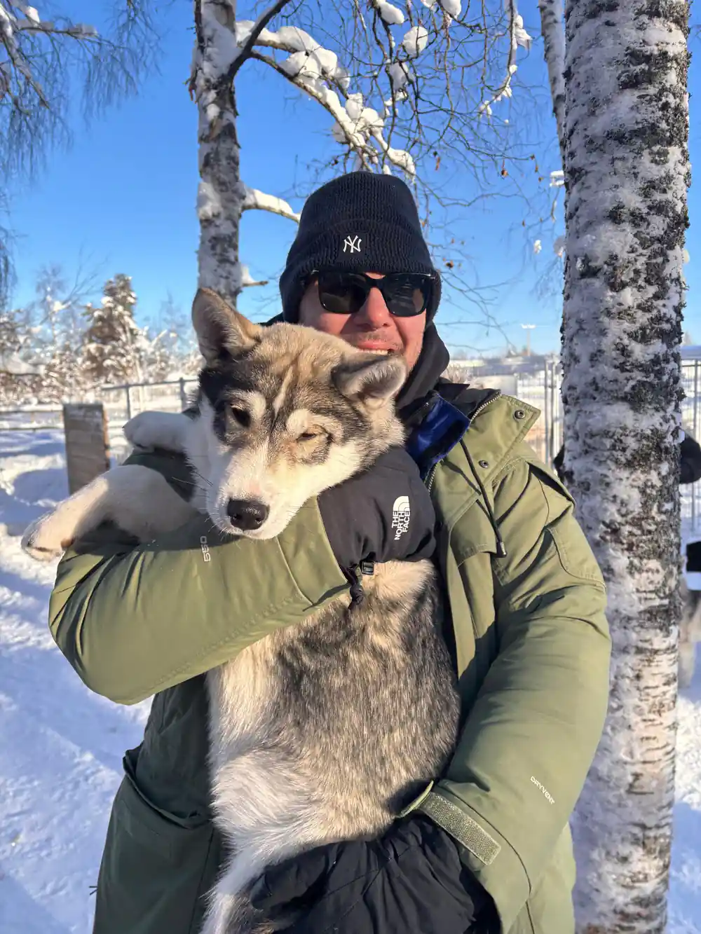 Man in sunglasses holding gray and white husky dog outdoors